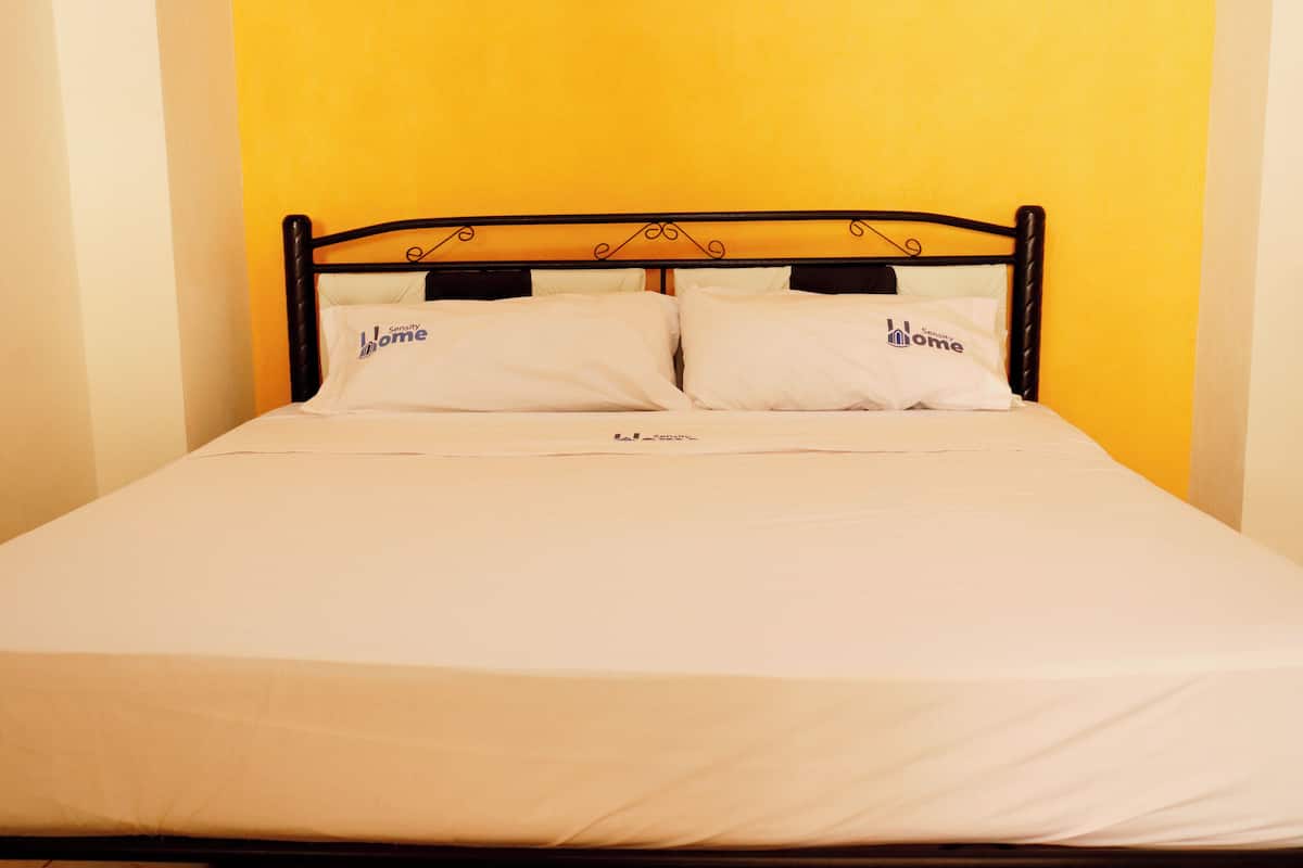 A bed with white bedding and two pillows, each with a logo, against a yellow wall.
