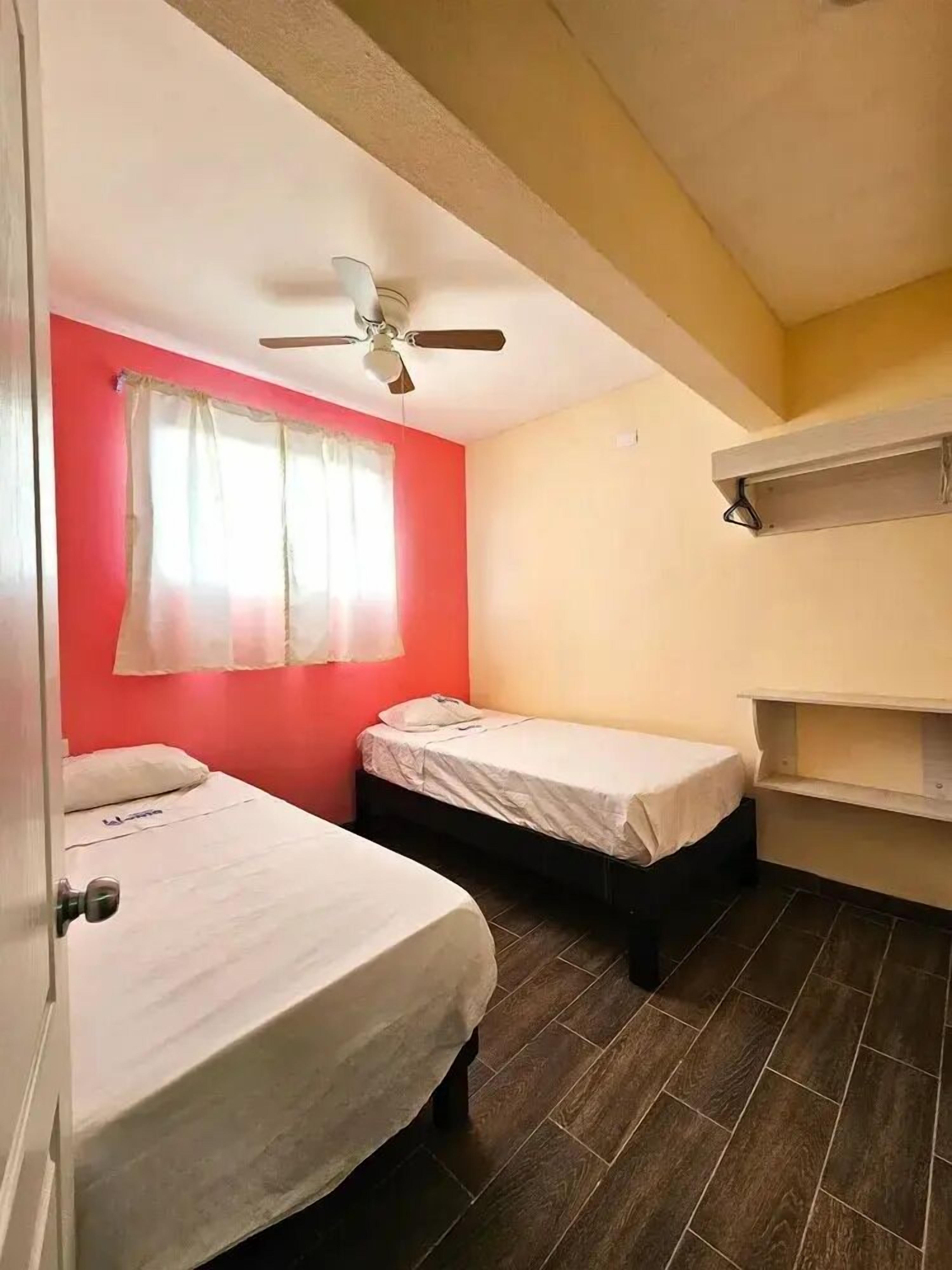 A room with two beds, a ceiling fan, a window with curtains, and a shelf unit.