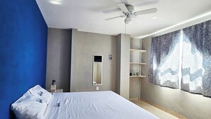 Un dormitorio moderno con una cama grande, un ventilador de techo, un estante montado en la pared y una ventana con cortinas estampadas.