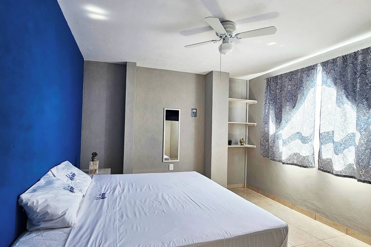 A modern bedroom with a large bed, a ceiling fan, a wall-mounted shelf, and a window with patterned curtains.