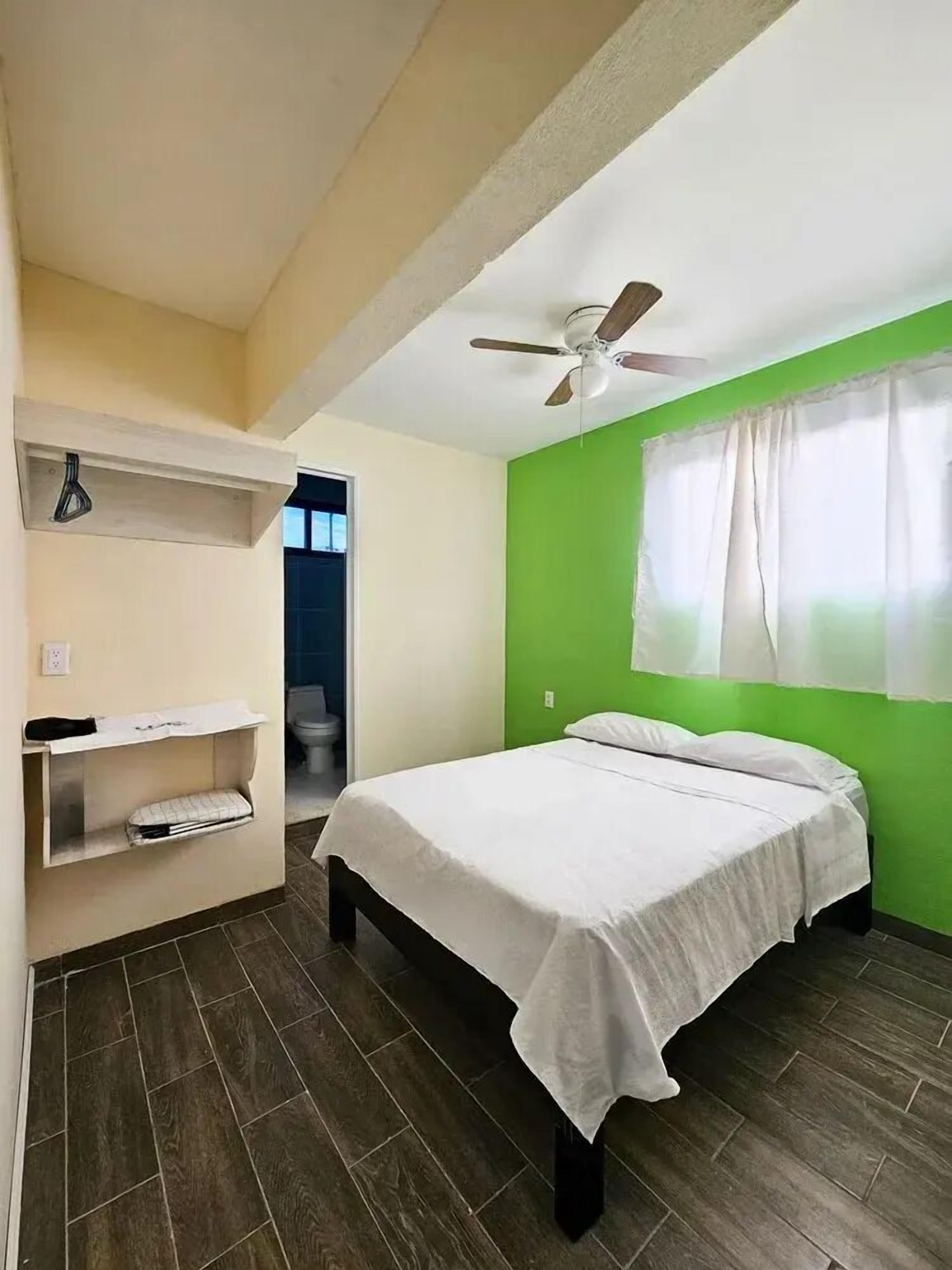 Une chambre à coucher comprenant un lit, un ventilateur de plafond, une fenêtre avec des rideaux et une porte donnant sur une salle de bain.
