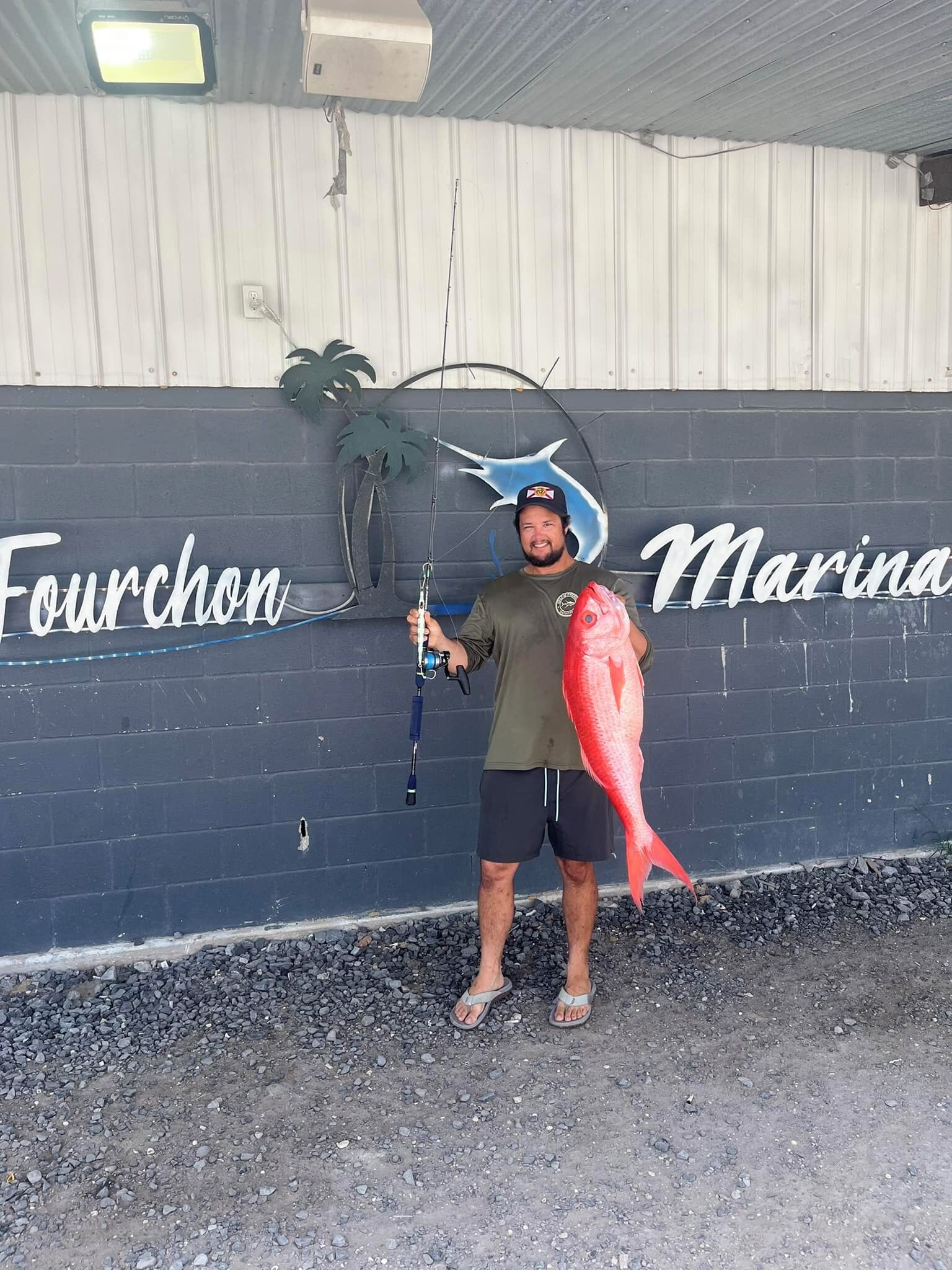 En mand holder en stor fisk foran en mur med teksten "Fourchon Marina".