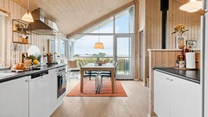 A modern kitchen with wooden cabinets, a dining area with a table and chairs, and large windows offering a view of the outdoors. - Cozy home in Lemvig with sauna (Lemvig)