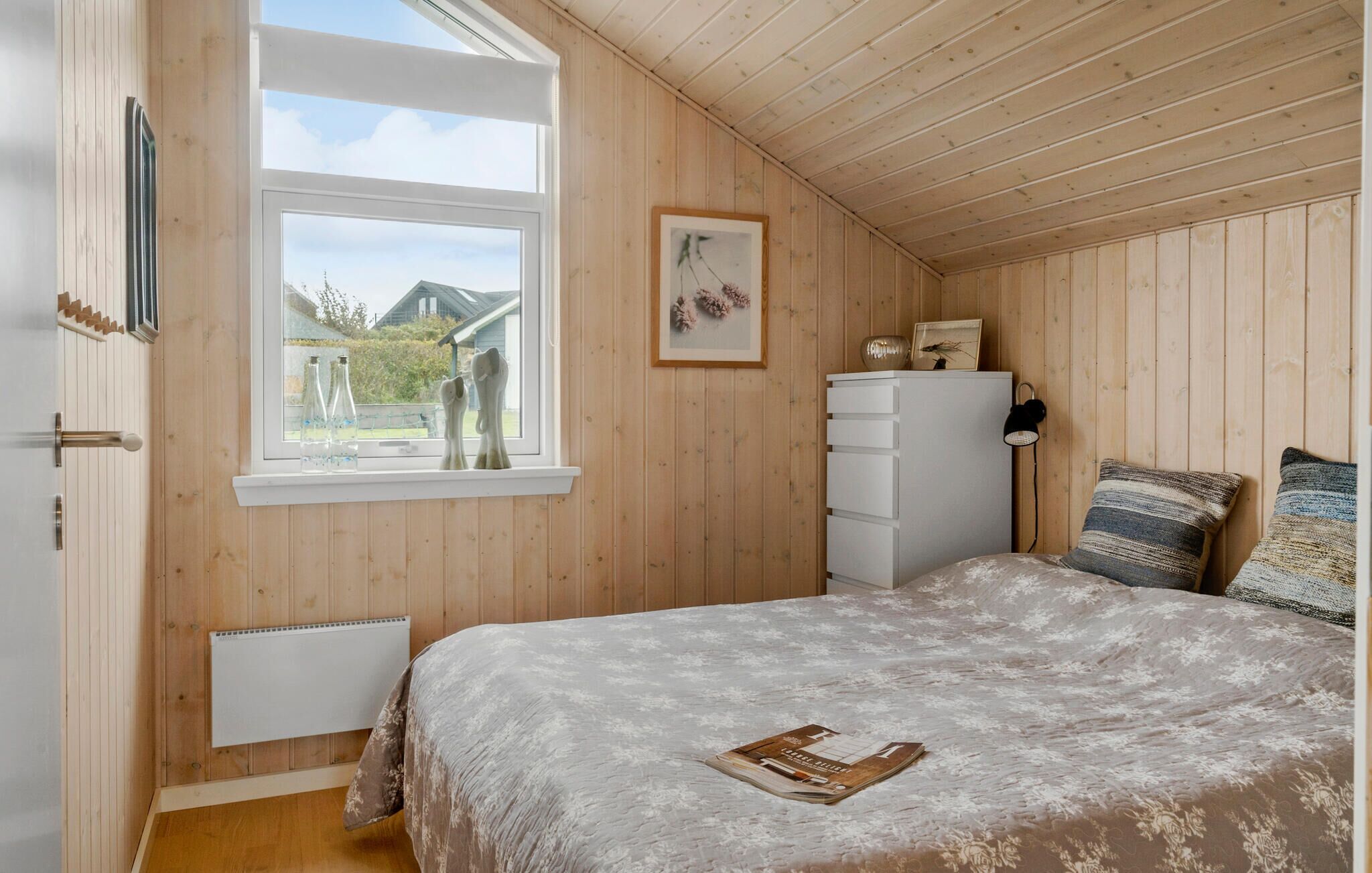 Un dormitorio con paredes de madera, un ventanal grande, una cama con colcha estampada, un aparador y un cuadro en la pared.