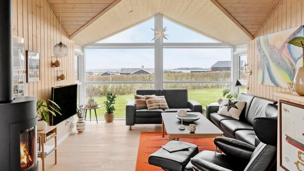 A cozy living room with a fireplace, comfortable seating, and large windows offering a view of the countryside. - Cozy home in Lemvig with sauna (Lemvig)