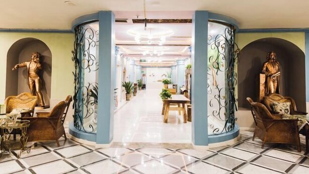 Eine Hotelhalle mit einem offenen zentralen Bereich, der von Säulen mit dekorativer Metallverzierung flankiert wird, einem Essbereich mit Tischen und Stühlen sowie einer Statue auf jeder Seite.