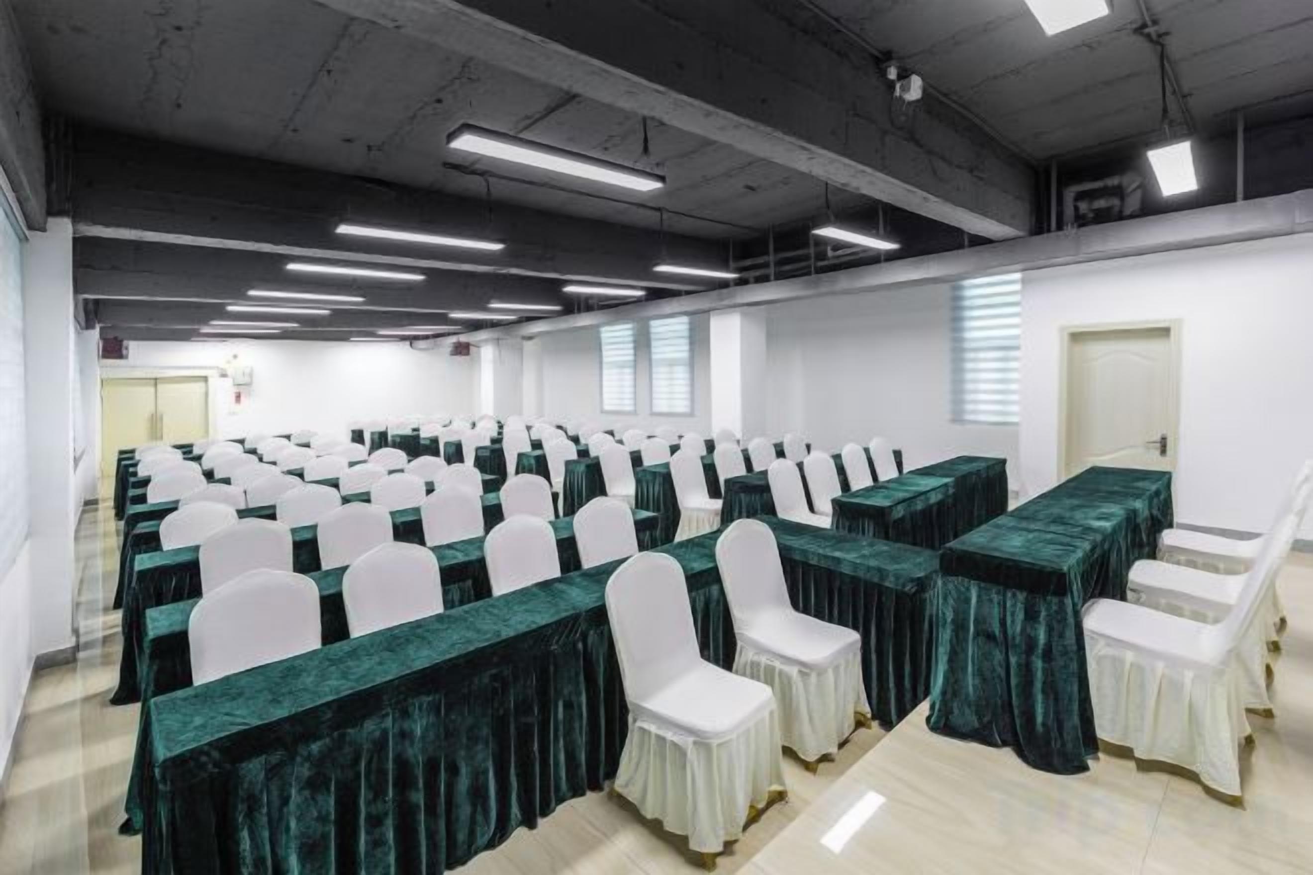 Un salón de conferencias con filas de mesas y sillas, un techo alto con iluminación industrial y una puerta al fondo.