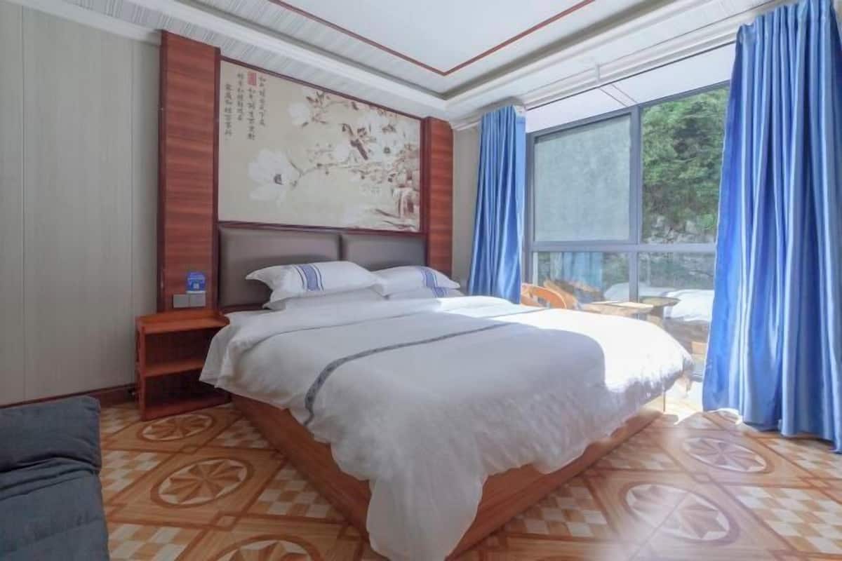 Een hotelkamer met een groot bed, een houten hoofdbord, een schilderij aan de muur, een tafeltje en uitzicht op groen door een raam.