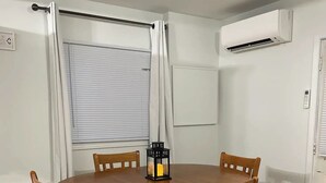 Un comedor con una mesa y sillas de madera, una ventana con persianas, un equipo de aire acondicionado y un aplique de pared.