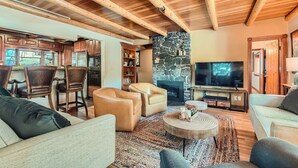 A spacious living room with a stone fireplace, wooden ceiling beams, and a television. - Historic Stone Mountain Lodge with Private Pond - convenient and secluded (Conifer)