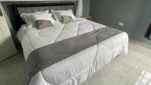 Una cama bien hecha con ropa de cama blanca y gris, un cabecero oscuro y una pared lisa con un interruptor de luz.