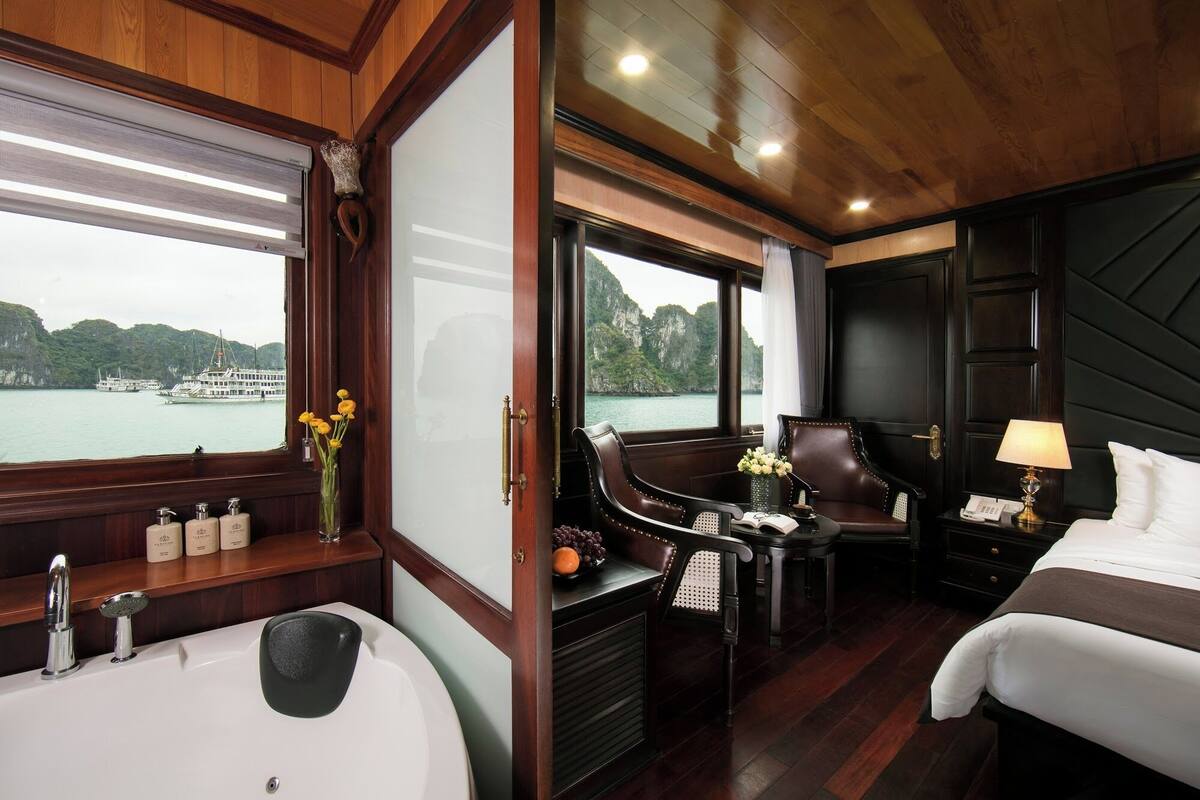 A room with a large window, a bathtub, a desk with a chair, a bed, and a nightstand with a lamp.
