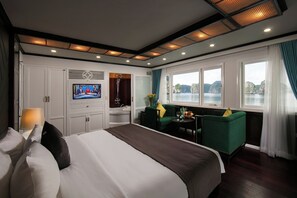 A hotel room with a large bed, a television, a seating area with a table, and a view of the water. - La Regina Royal Cruise (Ha Long)
