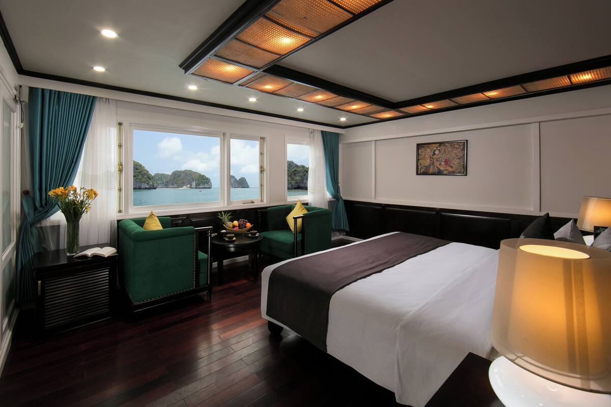 A hotel room with a bed, a desk, two chairs, a window with a view of the sea, and a painting on the wall.