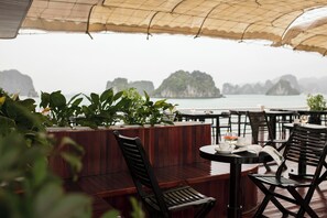 An outdoor seating area with wooden furniture and potted plants, overlooking a body of water with islands in the distance. - La Regina Royal Cruise (Ha Long)