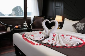 A hotel room with a bed adorned with a heart-shaped towel arrangement and rose petals, a view of the sea, a dark wooden headboard, and a nightstand with a lamp.