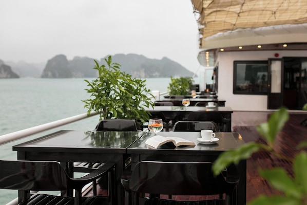 A table with an open book, a glass of water, and a cup on a boat deck with a view of the sea and distant mountains.