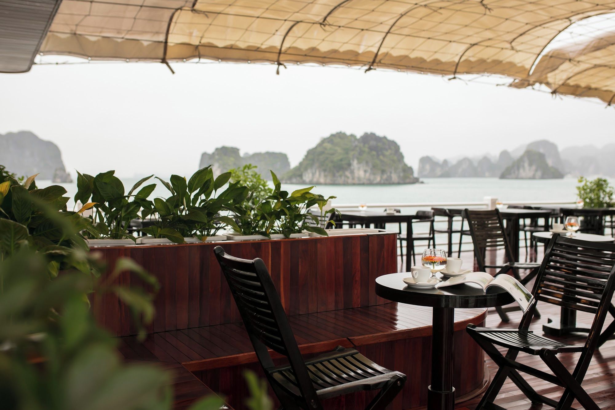 An outdoor seating area with wooden furniture and potted plants, overlooking a body of water with islands in the distance.
