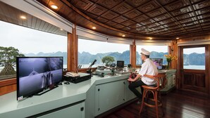 A control room with a large monitor, a steering wheel, and a view of mountains through the windows. - La Regina Royal Cruise (Ha Long)