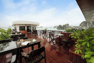 Un restaurante con mesas y sillas en una terraza de madera con vista a una masa de agua y acantilados rocosos.