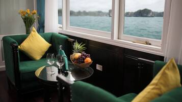 A room with a green sofa, a small table with a fruit bowl and wine glasses, and a view of the sea through large windows.