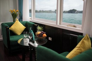 A room with a green sofa, a small table with a fruit bowl and wine glasses, and a view of the sea through large windows. - La Regina Royal Cruise (Ha Long)
