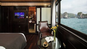 A room with a TV, a table with a book and a cup, a chair, and a view of the sea. - La Regina Royal Cruise (Ha Long)