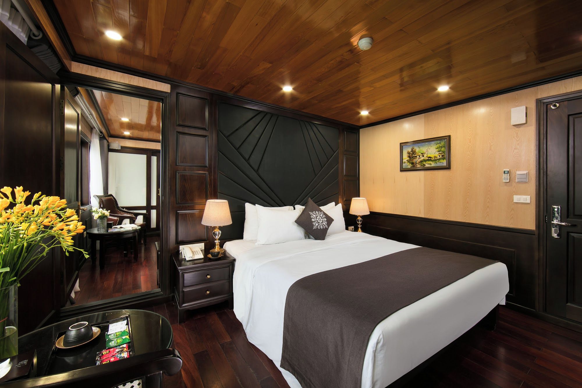 A hotel room with a wooden ceiling, a bed with white bedding, a nightstand with a lamp, a desk with a telephone, and a painting on the wall.