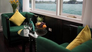 A room with a green sofa, a small table with a fruit bowl and wine glasses, and a view of the sea through large windows. - La Regina Royal Cruise (Ha Long)