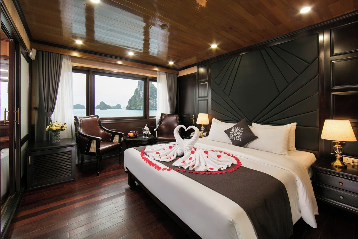 Habitación de hotel con cama, dos sillas, una mesita, una lámpara y vista al mar.