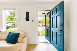 A room with a beige sofa, teal cushions, a teal door, and a view of a street with houses.