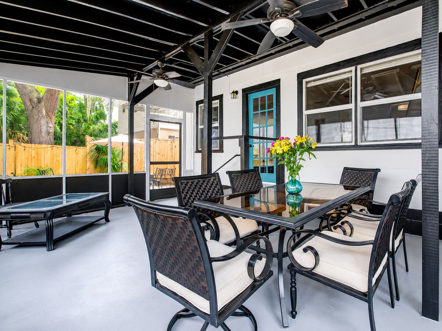Un patio al aire libre con una mesa y sillas de vidrio, un ventilador de techo y vista a una cerca de madera y vegetación.