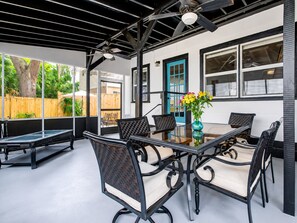 Une terrasse extérieure avec une table et des chaises en verre, un ventilateur de plafond et une vue sur une clôture en bois et de la verdure.