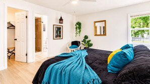 A bedroom with a bed, a hanging plant, a chair, a window, and a door leading to another room.