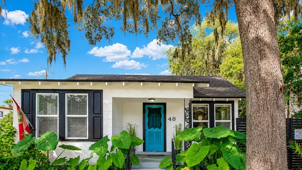 A single-story house with a blue door, number 48, surrounded by large green plants and a tree.
