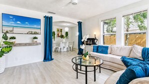 A spacious living room with a large window, a ceiling fan, a dining area with white chairs, and a sofa with blue cushions.