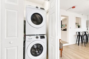 Un cuarto de lavandería con dos lavadoras apiladas, una zona de comedor con una mesa y sillas, y una cocina al fondo.
