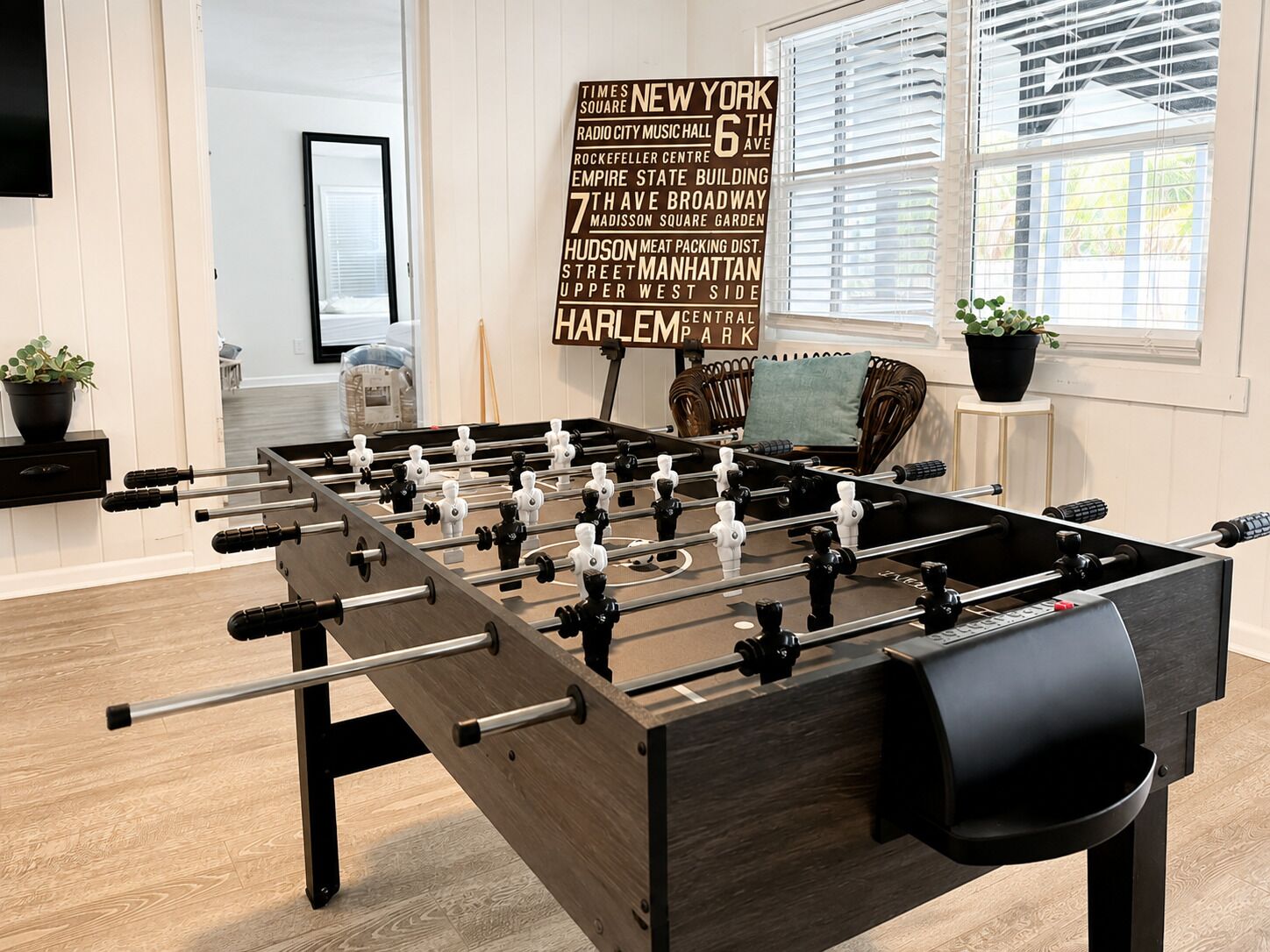 Una mesa de fútbol de mesa en una habitación con un ventanal grande, un cartel enmarcado con puntos de referencia de Nueva York y una silla de mimbre.