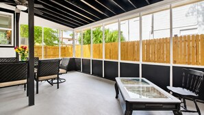 A covered outdoor seating area with a glass-top table, black chairs, and a wooden fence in the background.
