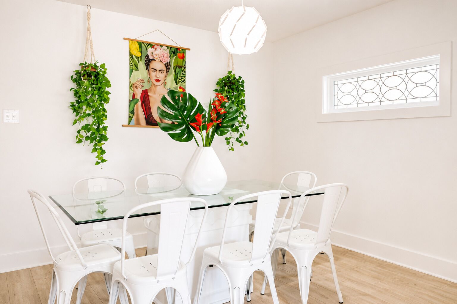 Un coin repas avec une table en verre, des chaises blanches, un vase blanc contenant des fleurs rouges et un tableau accroché au mur.