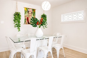 Un comedor con una mesa de vidrio, sillas blancas, un jarrón blanco con flores rojas y un cuadro colgado en la pared.