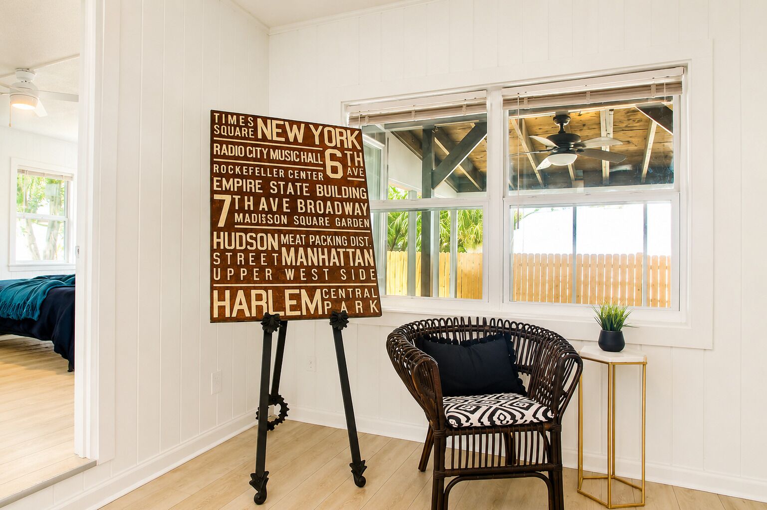 Una habitación con un cartel grande que enumera lugares emblemáticos de Nueva York, una silla de mimbre, una mesita con una planta y una ventana con vista a una valla de madera.