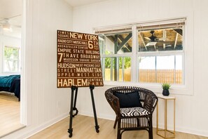 Une pièce avec une grande affiche présentant les principaux monuments de New York, une chaise en osier, une petite table avec une plante et une fenêtre donnant sur une clôture en bois.