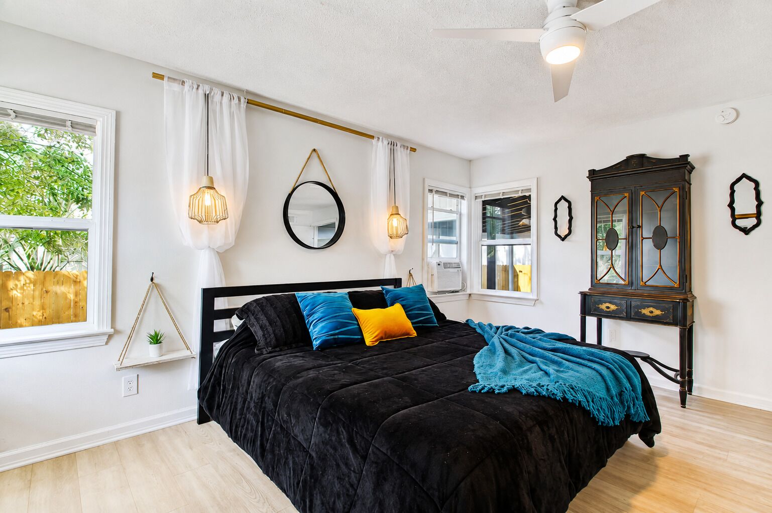 Une chambre à coucher avec un lit noir, des murs blancs, un ventilateur de plafond et une fenêtre avec des rideaux.