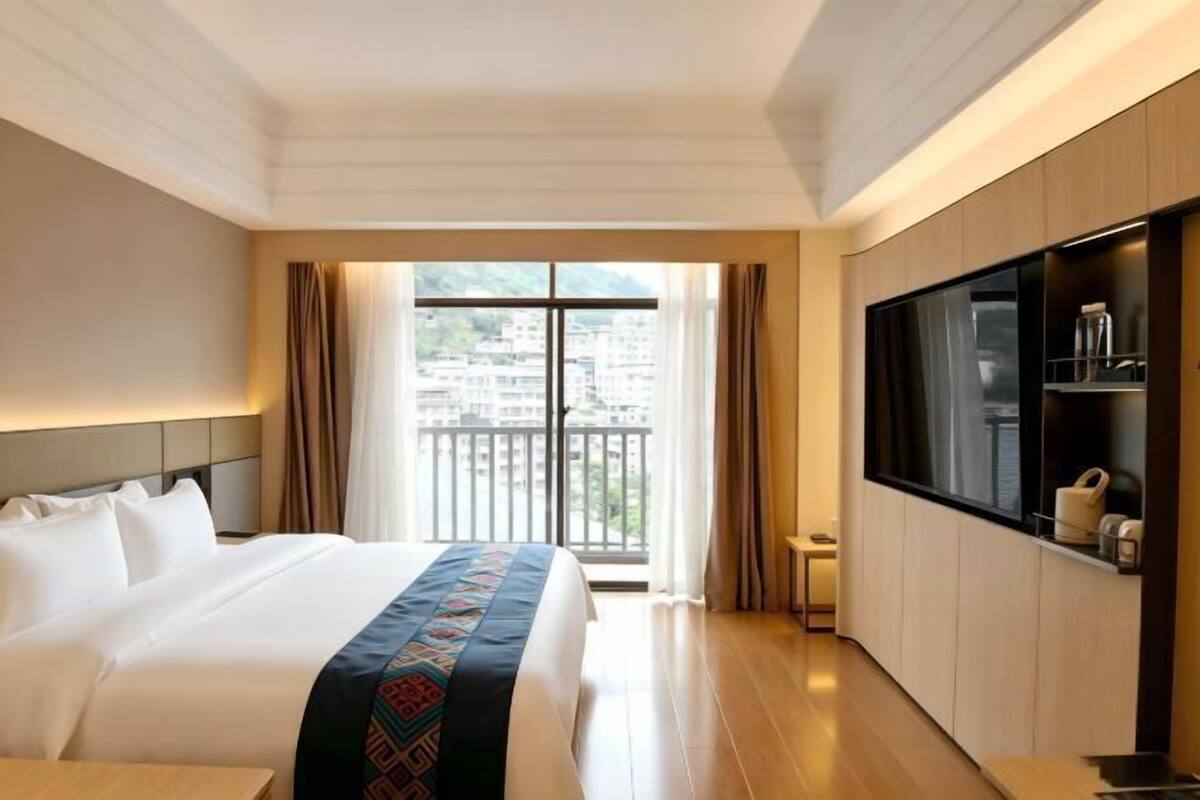 Habitación de hotel con cama, televisor, una mesita y balcón con vistas a edificios.