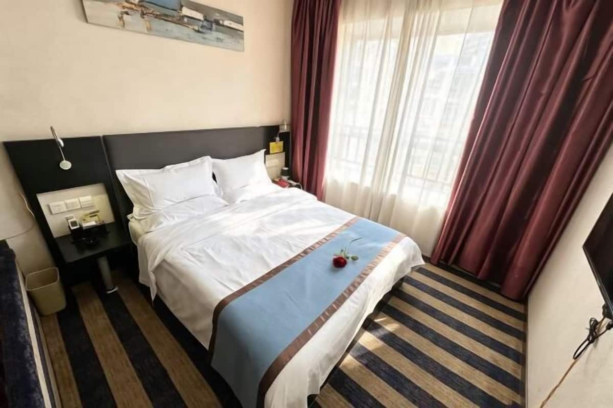 A hotel room with a bed, a nightstand, a lamp, a window with curtains, and a wall-mounted artwork.