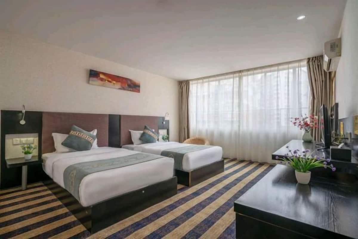 A hotel room with two beds, a desk, a chair, a potted plant, and a window with curtains.