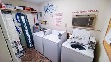 Une buanderie équipée de deux machines à laver, d’un sèche-linge et d’un panneau indiquant les heures d’ouverture et les règles d’utilisation.