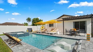 A swimming pool with lounge chairs and umbrellas, a hot tub, and a patio area with a table and chairs.