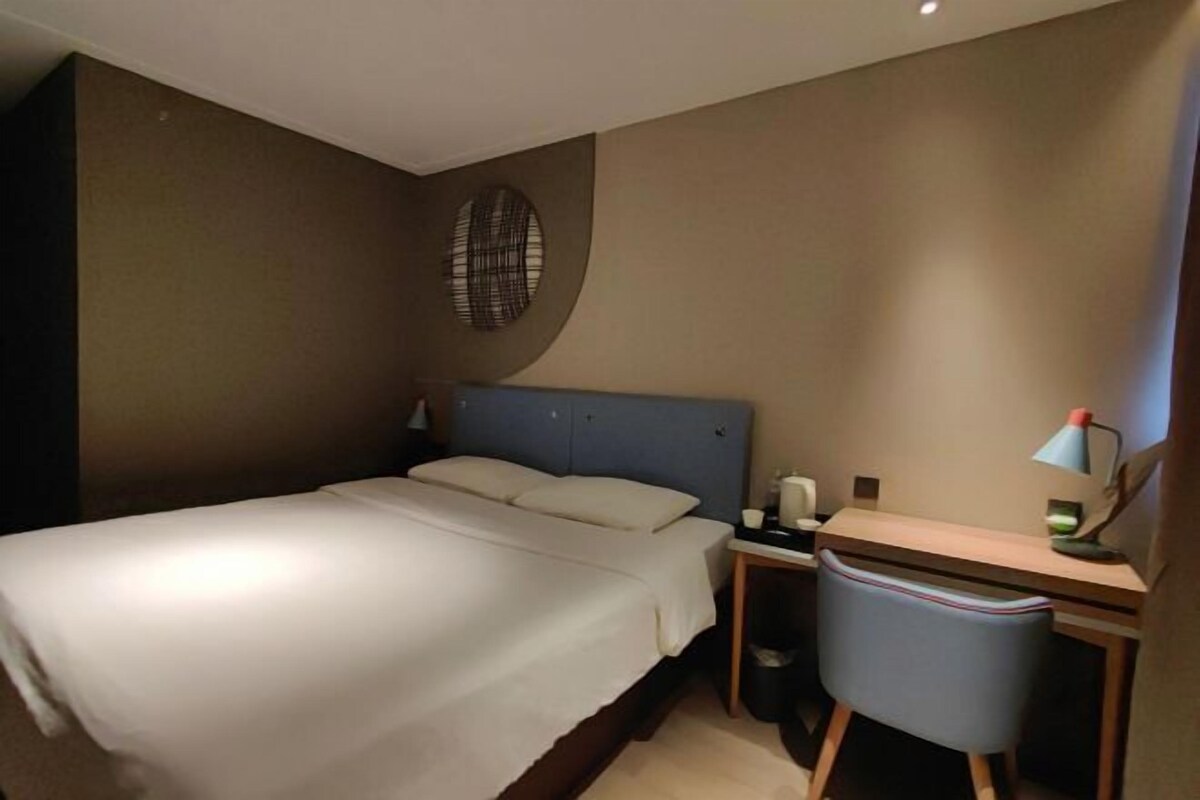 A hotel room with a bed, desk, chair, lamp, and waste bin.